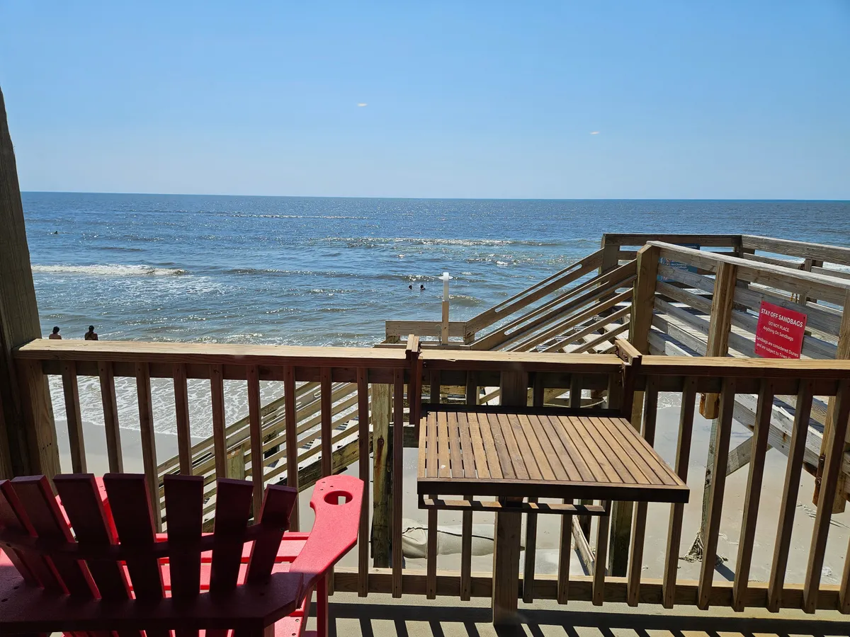 A serene view from the private balcony, featuring a red Adirondack chair, wooden railing, and panoramic ocean scenery with distant surfers. The inviting setup suggests enjoying a drink, sunbathing, or watching the sunrise and sunset while immersed in the sound of waves.