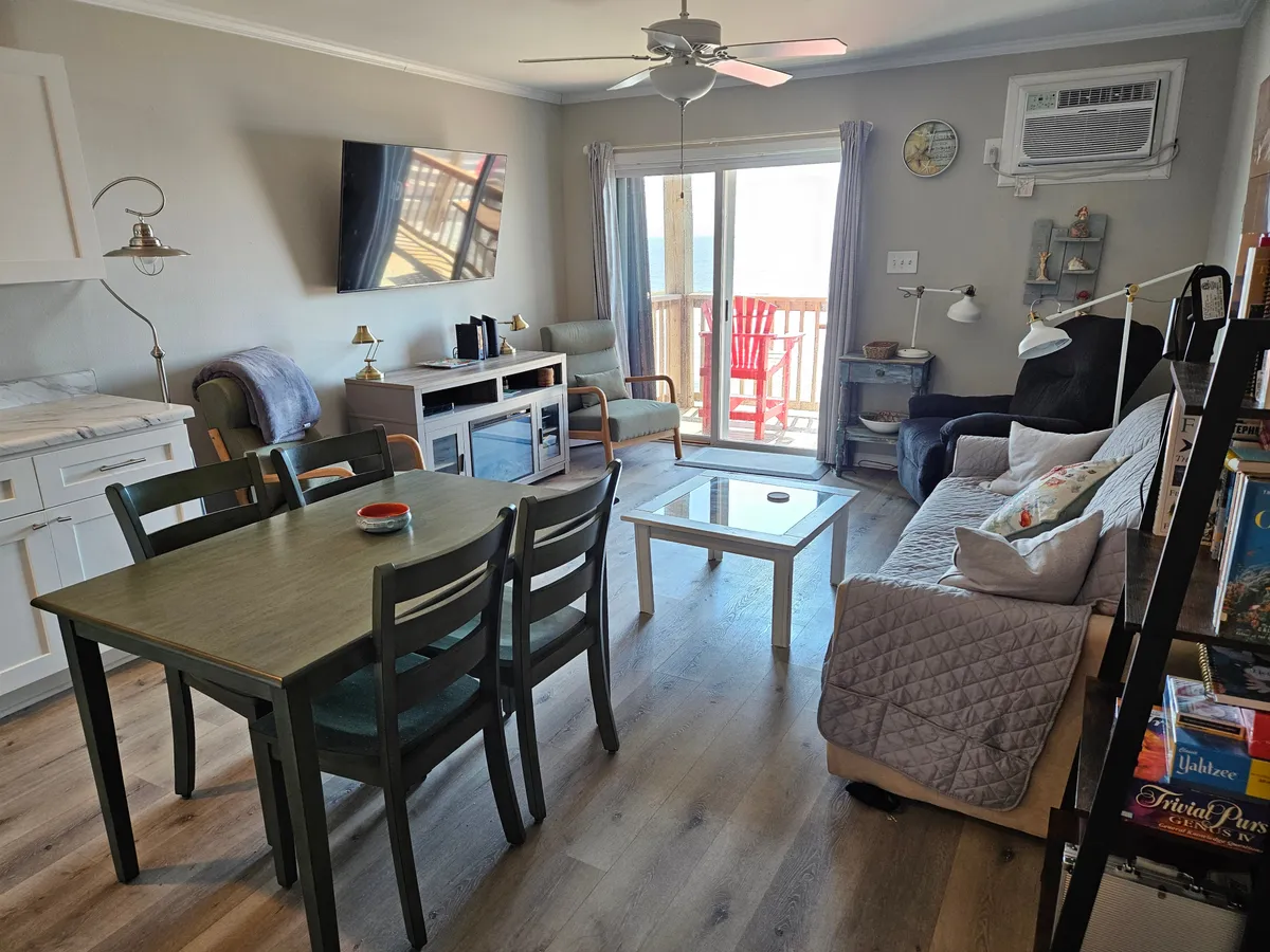 The image captures a welcoming living and dining area with a table, a plush sofa adorned with decorative pillows, and a smart TV. Red chairs are visible on the private balcony just outside, enabling a clear visual link between the interior and the outdoor space, with glimpses of the ocean beyond.