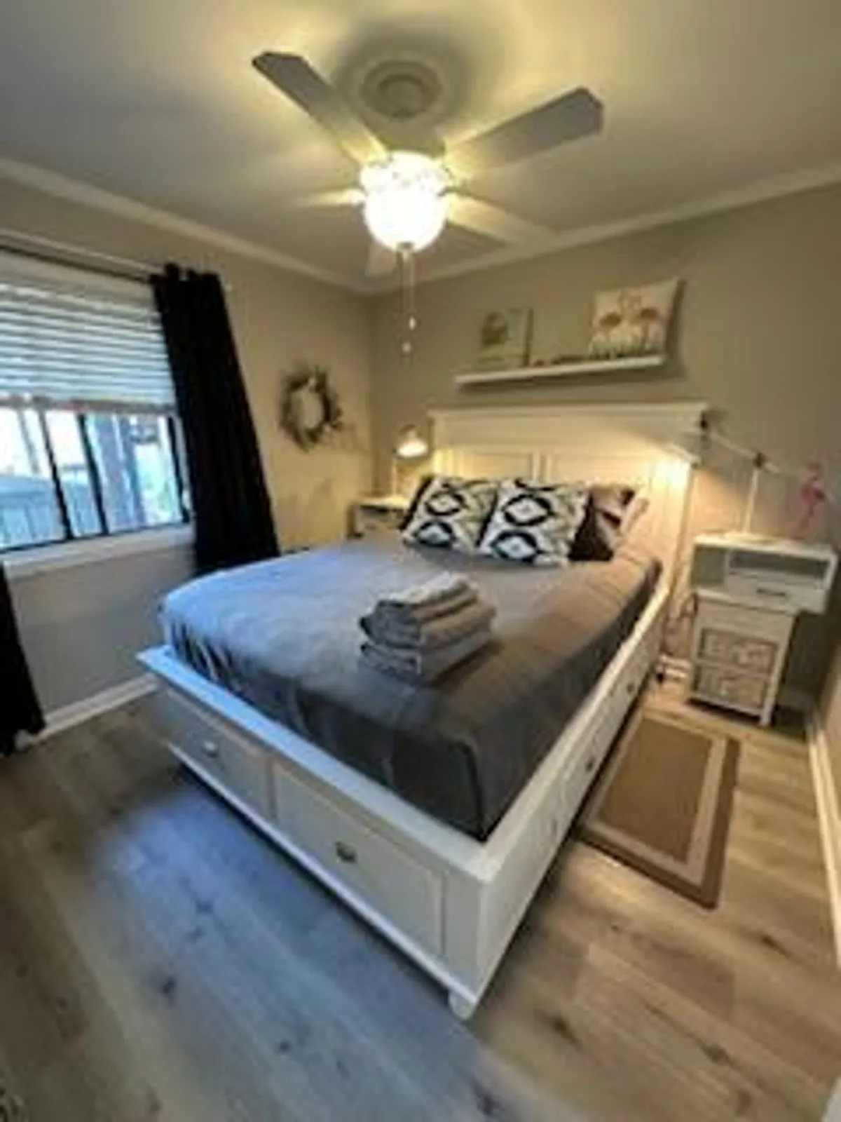 A cozy bedroom scene featuring a neatly made bed, nightstand, and ceiling fan. Natural light filters in, enhancing the inviting mood ideal for relaxation and recharging after a day by the sea.