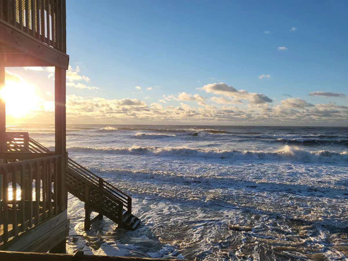 A stunning sunrise bathes the ocean in warm light, with gentle waves and wooden stairs leading down to the sandy beach, inviting guests to stroll along the shore.
