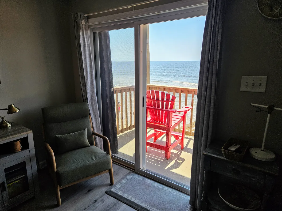 A comfortable red chair sits on the private deck, offering an inviting spot to relax and enjoy unobstructed ocean views. Inside, another cozy chair is visible through the glass, blending indoor comfort with the outdoor serenity of the beachfront.
