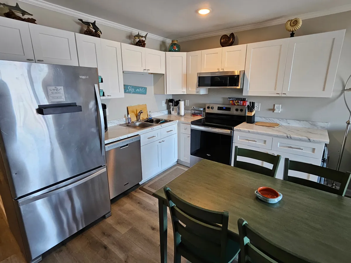 This image showcases a bright, functional kitchen with a stainless steel refrigerator, white cabinetry, and a lively green dining table. Decorative items on the shelves add a personalized touch while the space is clearly set up for cooking, dining, and entertaining. The layout and finishes underscore the gourmet and inviting nature of the kitchen.