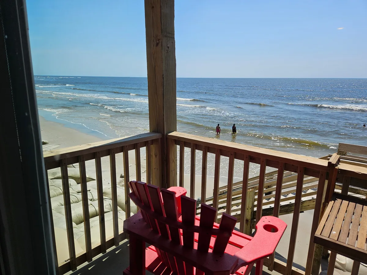The image features a cozy red Adirondack chair on a private balcony, overlooking gentle ocean waves and a sandy beach dotted with people relaxing, swimming, and beachcombing. The tranquil scene highlights the easy access from the condo directly onto the beach, perfect for sunrise views, building sandcastles, and sunset strolls along the shore.