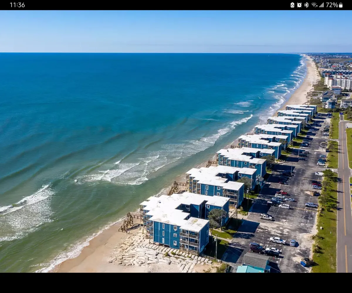 A sweeping exterior view of beachfront buildings with the ocean waves rolling ashore. The foreground includes a parking area, with the shoreline just steps away, inviting for beach days. The scene captures families and groups enjoying leisurely walks and activities right by the water, while nearby restaurants and shops remain within easy reach.