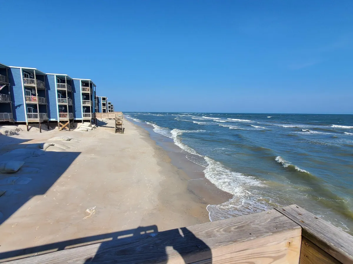 A wide exterior shot showing the beachfront condominium buildings, a wooden walkway leading toward the sand, and gentle waves lapping the shore. The setting is tranquil and welcoming, emphasizing easy access to the beach and the proximity of living spaces to the ocean.