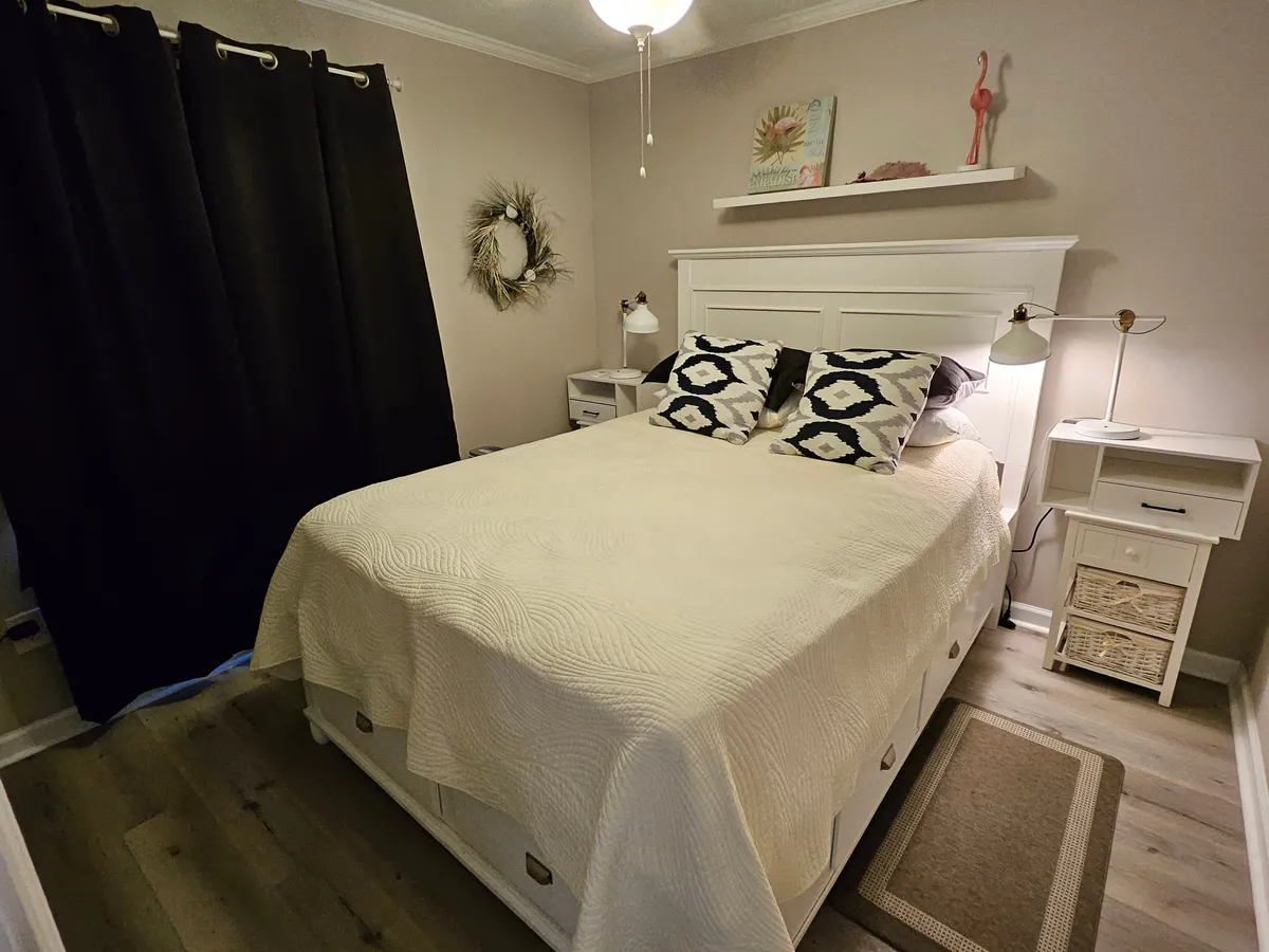 A serene bedroom with a comfortable bed dressed in premium linens, flanked by nightstands and accented with blackout curtains and tasteful wall art. Soft lighting and a decorative wreath add to the calm, inviting atmosphere.