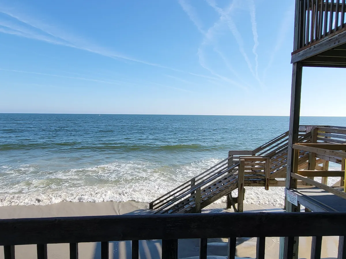 A view of the ocean and sandy beach with stairs leading down, inviting guests to explore the shoreline. The inviting scene highlights the beauty of the ocean and the accessibility of the beach area for breaks or a change of workspace scenery.